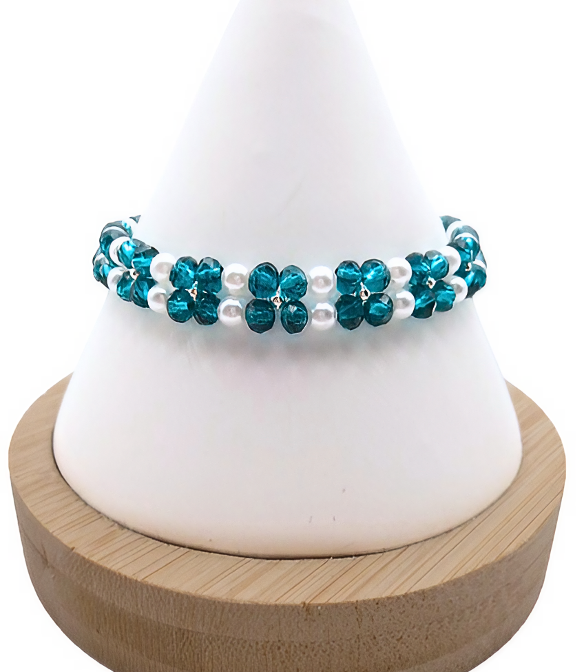 Handmade bracelet featuring teal faceted crystals arranged in a repeating four-petal flower pattern with white pearl centres and borders, and silver seed bead accents, displayed on a white cone bracelet stand with a wooden base against a white background.