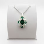 A pendant made with faceted emerald green crystal surrounded by silver beads and white pearls, shown with a silver bail and a silver chain, displayed on a white stand.