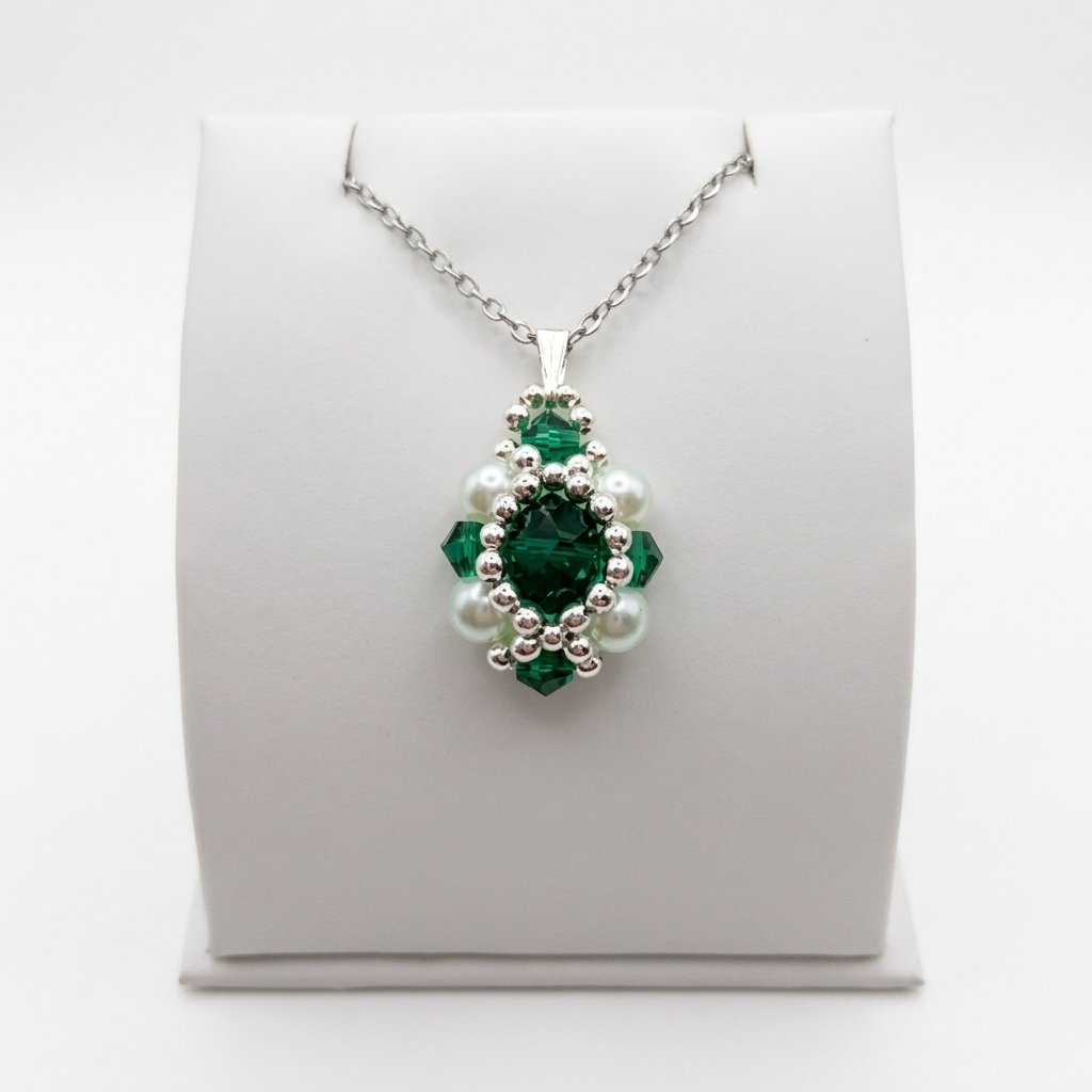 emerald-crystal-pearl-pendant-display A pendant made with faceted emerald green crystal surrounded by silver beads and white pearls, shown with a silver bail and a silver chain, displayed on a white stand.