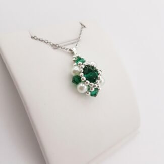 A side view of a pendant made with faceted emerald green crystal surrounded by silver beads and white pearls, shown with a silver bail and a silver chain, displayed on a white stand.