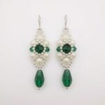 Handcrafted drop earrings made with deep emerald green crystals, white pearl beads, and silver accents hanging against a neutral background.