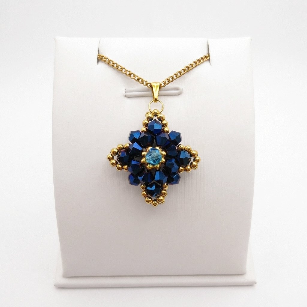 navy-blue-crystal-pendant-display Navy blue crystal pendant with metallic blue crystals, aquamarine-coloured centre crystal, gold-coloured bead accents and matching chain. Shown on a display stand.
