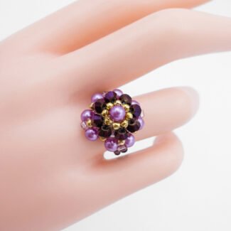 A unique ring featuring a beautiful cluster of purple pearls and darker metallic purple crystals, accented with gold beads. It is shown on a mannequin hand for scale and a white background for detail.