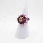 A side view of the beaded ring with dark metallic purple crystals, lilac pearls and gold beads in a flower design, displayed on a white ring cone