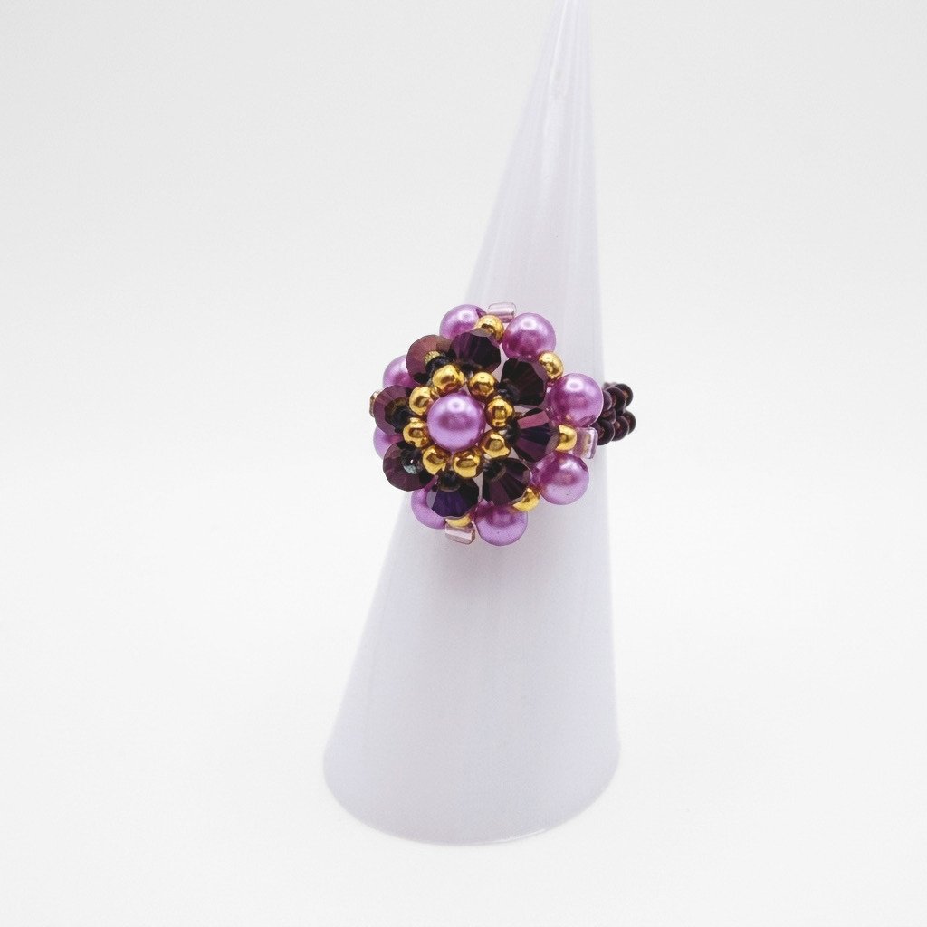 purple-crystal-pearl-ring-side-view A side view of the beaded ring with dark metallic purple crystals, lilac pearls and gold beads in a flower design, displayed on a white ring cone