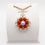 Front view of flower-design pendant featuring off-white pearls and faceted red crystals. The pendant has a matching gold bail and a gold chain, shown on a display stand.