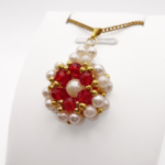 Side view of flower-design pendant featuring off-white pearls and faceted red crystals. The pendant has a matching gold bail and a gold chain, shown on a display stand.