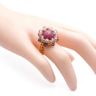 Close-up of a handcrafted ring featuring a tourmaline stone. The central stone is surrounded by a halo of gold and faceted purple beads, all set on an intricate beaded gold band. The ring is displayed on a mannequin hand against a clean white background.