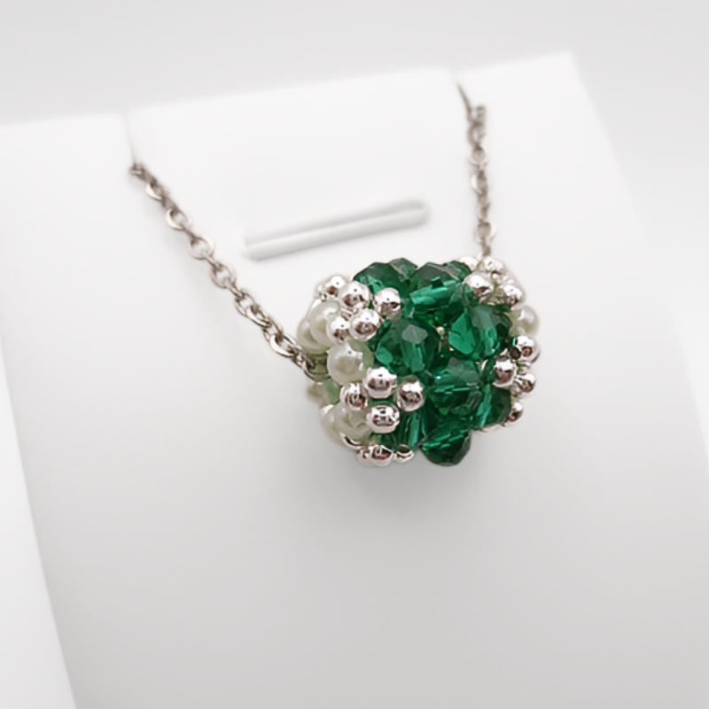 barrel-crystal-pendant-green Silver-tone chain necklace with an intricate woven beaded barrel-shaped pendant made of emerald green crystals and white pearls, shown on a display stand against a white background.