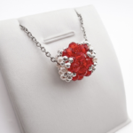 Silver-tone chain necklace with an intricate woven beaded barrel-shaped pendant made of red crystals and white pearls, shown on a display stand against a white background.